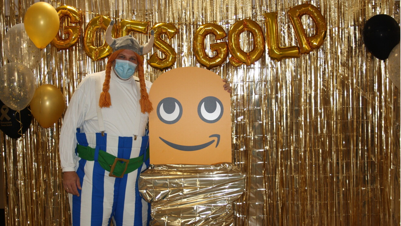 Ein verkleideter Mitarbeiter steht vor einer "Amazon Goes Gold"-Wand.