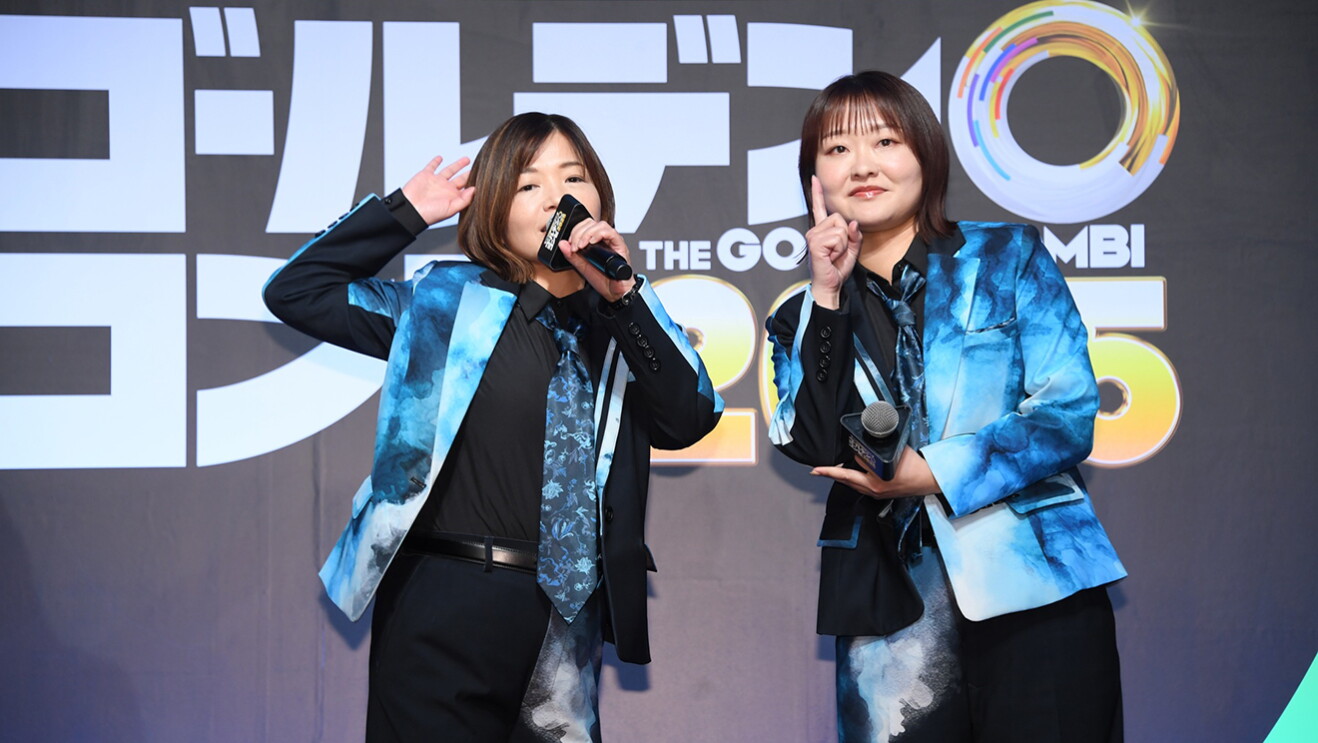 黒地にピンクと黄緑の曲線が入った背景の前で、黒と水色のマーブル模様のおそろいのスーツを着た2人の女性がマイクを持って立ち、おどけたポーズを取っている。