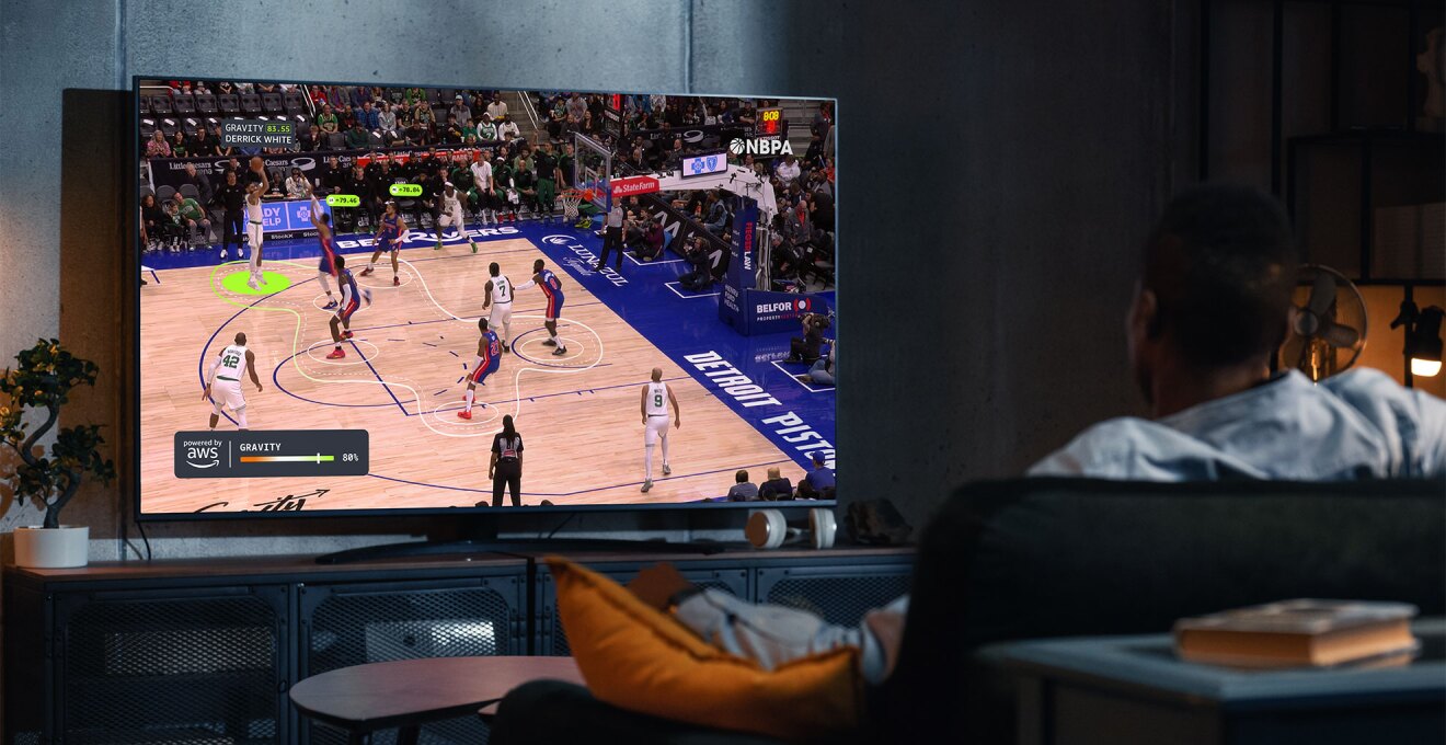 薄暗いリビングでソファに腰掛けてテレビを見る人物の後ろ姿。テレビにはバスケットボールの試合の様子が映る。画面にはハイライトや曲線の表示され、試合がデータ分析されていることがわかる。