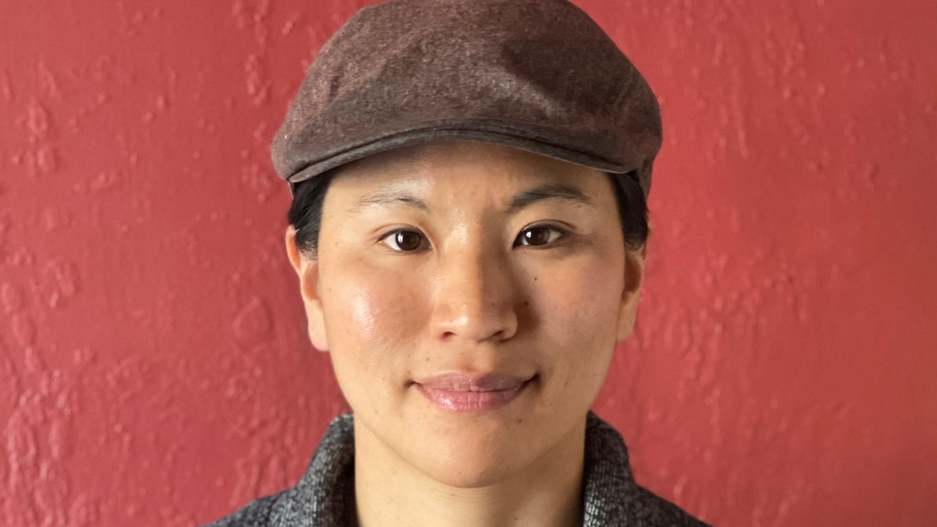 A woman wearing a newsboy cap, henley shirt, sweater, and necklace sits in front of a red wall.