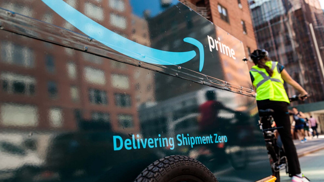 An Amazon delivery person riding a bicycle, with a bike trailer behind it. The trailer has "Delivering Shipment Zero" printed on it. 
