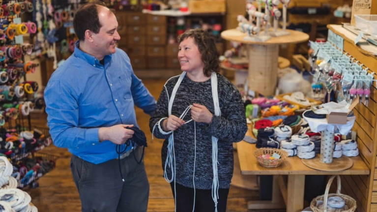 Two people standing and smiling inside Mystic Knotwork's brick-and-mortar store, one holding a handcrafted rope product with colorful nautical items on display.