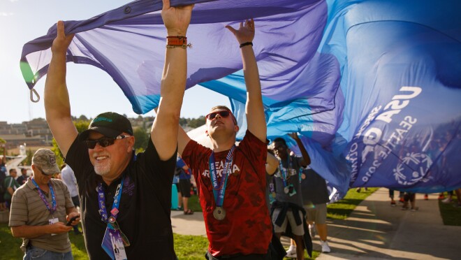 男运动员携带特殊奥运会旗仪式。运动员正拿着国旗在外缘,而带着它走向目的地。运动员在前台微笑,双手拿着国旗。第二个运动员正在向上支撑国旗,而其他运动员身后。前两个运动员背后的标志延伸了几英尺。事件参与者观看,在后台。