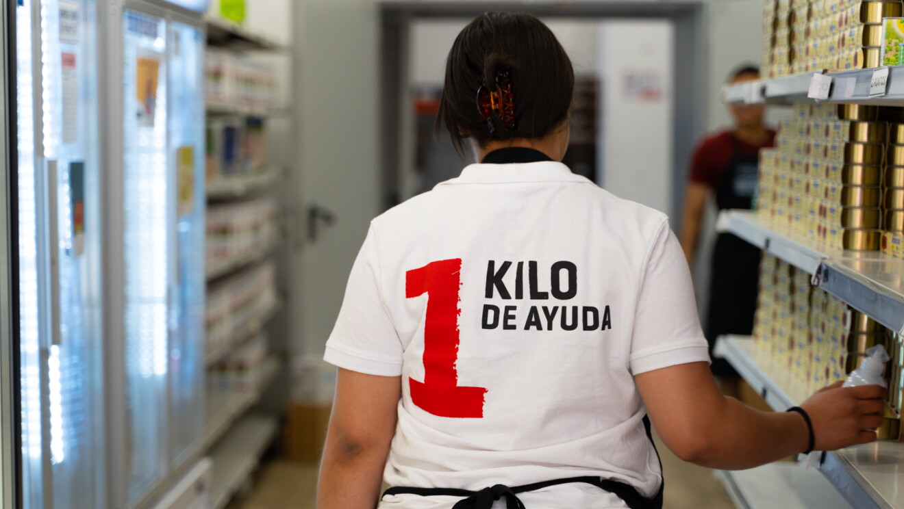 Una persona que parece una mujer, solo se le ve la espelda. Lleva un polo de color blanco con el 1 en números y de color rojo y después el texto: kilo de ayuda. Ella está agarrando algo con la ma no derecha ya los lados se ven dos estanterías, una con productos enlatados y las otras una parte con tetra bricks de leche y neveras frigoríficas. 