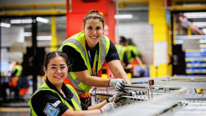 Dentro de un centro logístico, están dos mujeres arreglando una cadena metálica de transporte de paquetes. Las dos mujeres tiene el pelo oscuro largo y recogido en una cola. Las dos van en un camiseta negra de manga corta y un chaleco amarillo. Están las dos sonriendo , mirando a cámara, con las cuatro manos y agarrando algo y una de ellas agarra un destornillador.