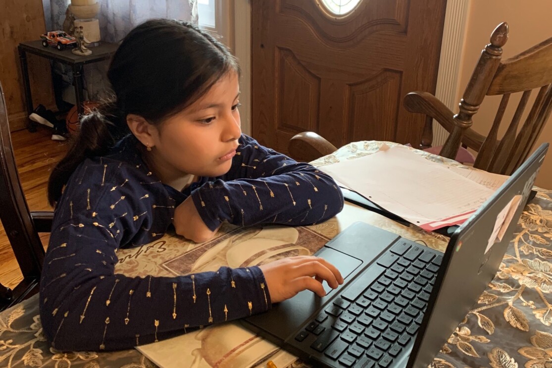 A girl sits a dining room table with a tablecloth and works on a laptop computer.