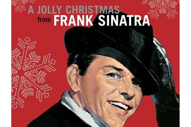 Frank Sinatra wears a suit and white button down, with a rimmed hat on his head, hair slicked, twinkle in his eye. His black gloved hand reaches up to tip his hat. He smiles toward the camera. The background is red, with large illustrated snowflakes falling around him.
