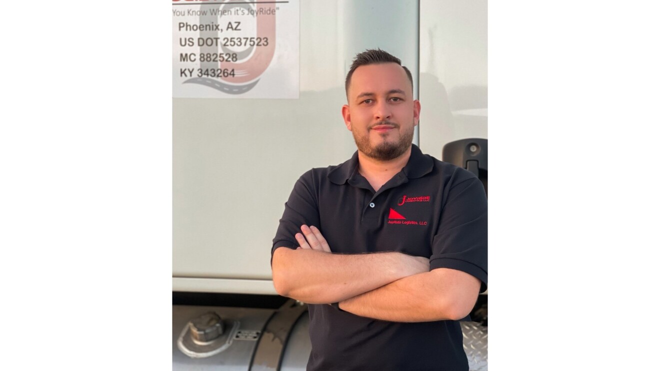 Adis Danan poses for the camera as he stands in front of his semi-truck. He stands with his arms crossed and wears a black collared T-Shirt.