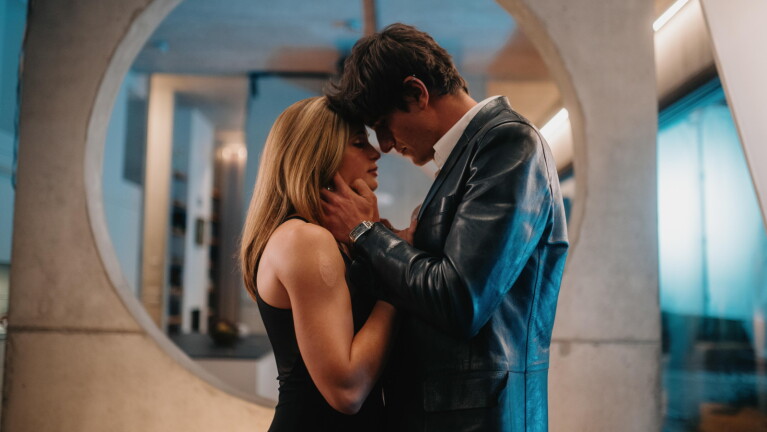A man and woman stand close together in an embrace, foreheads touching, in a modern interior lit with warm and cool blue tones.