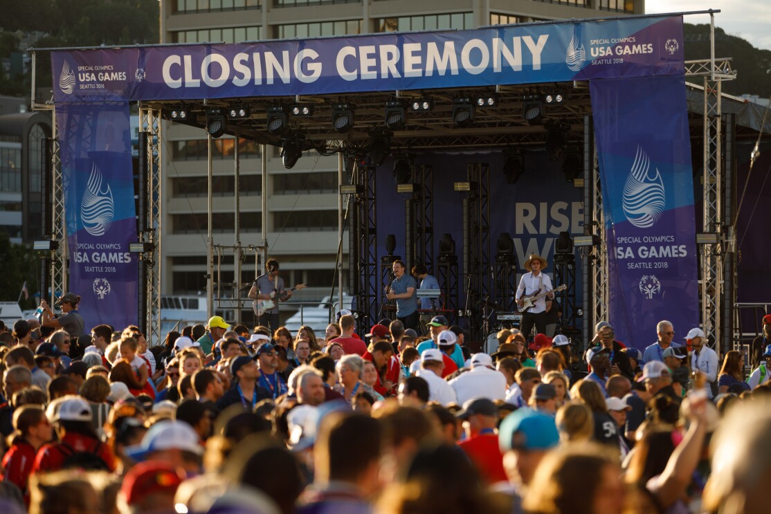 The 2018 Special Olympics USA Games closing ceremony stage. Seattle band "Hey Marseilles" performs (in the photo, a singer, bass player, keyboardist and electric guitarist), with a crowd of individuals in front of the stage.