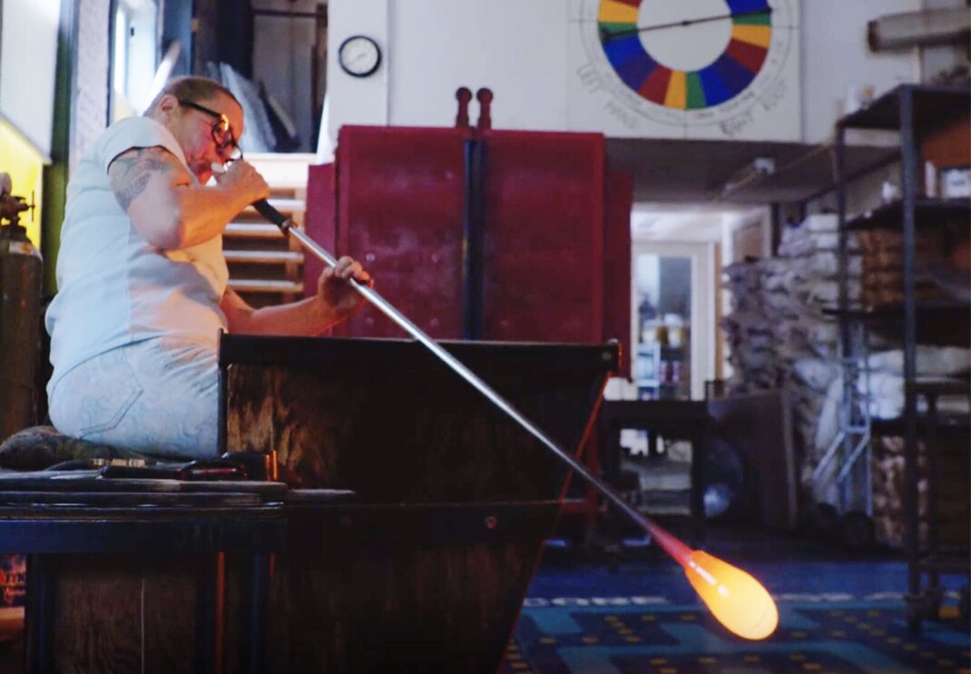 Dehanna Jones blows into a long tool to shape the glass at the end of the tool.