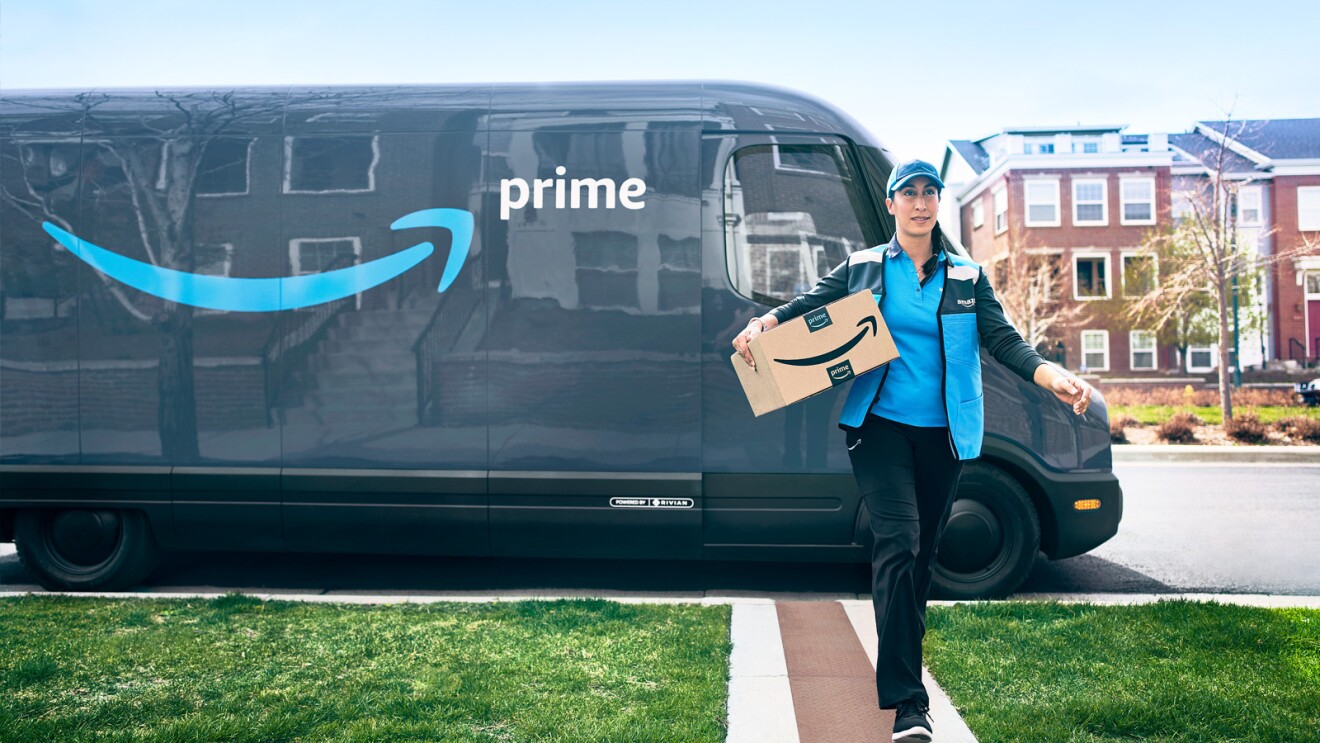 An Amazon delivery driver carries a package with a Rivian delivery vehicle parked on the street behind them.