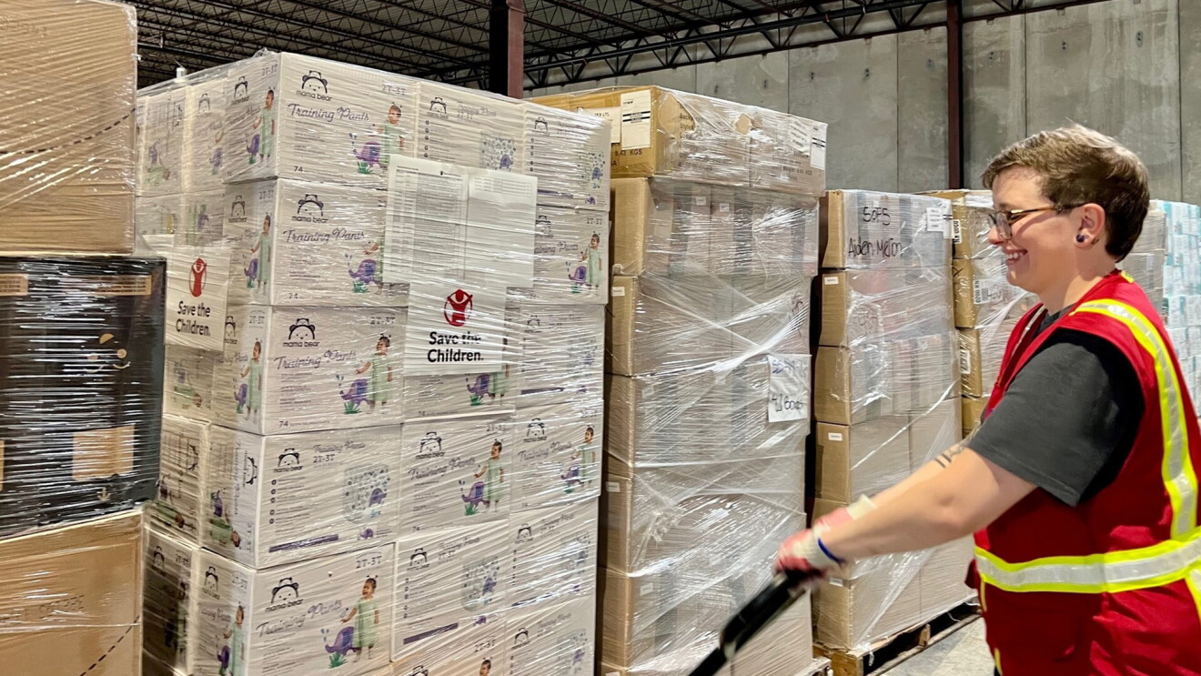 A volunteer wears a safety vest and smiles as they move a pallet of items being donated with a machine.