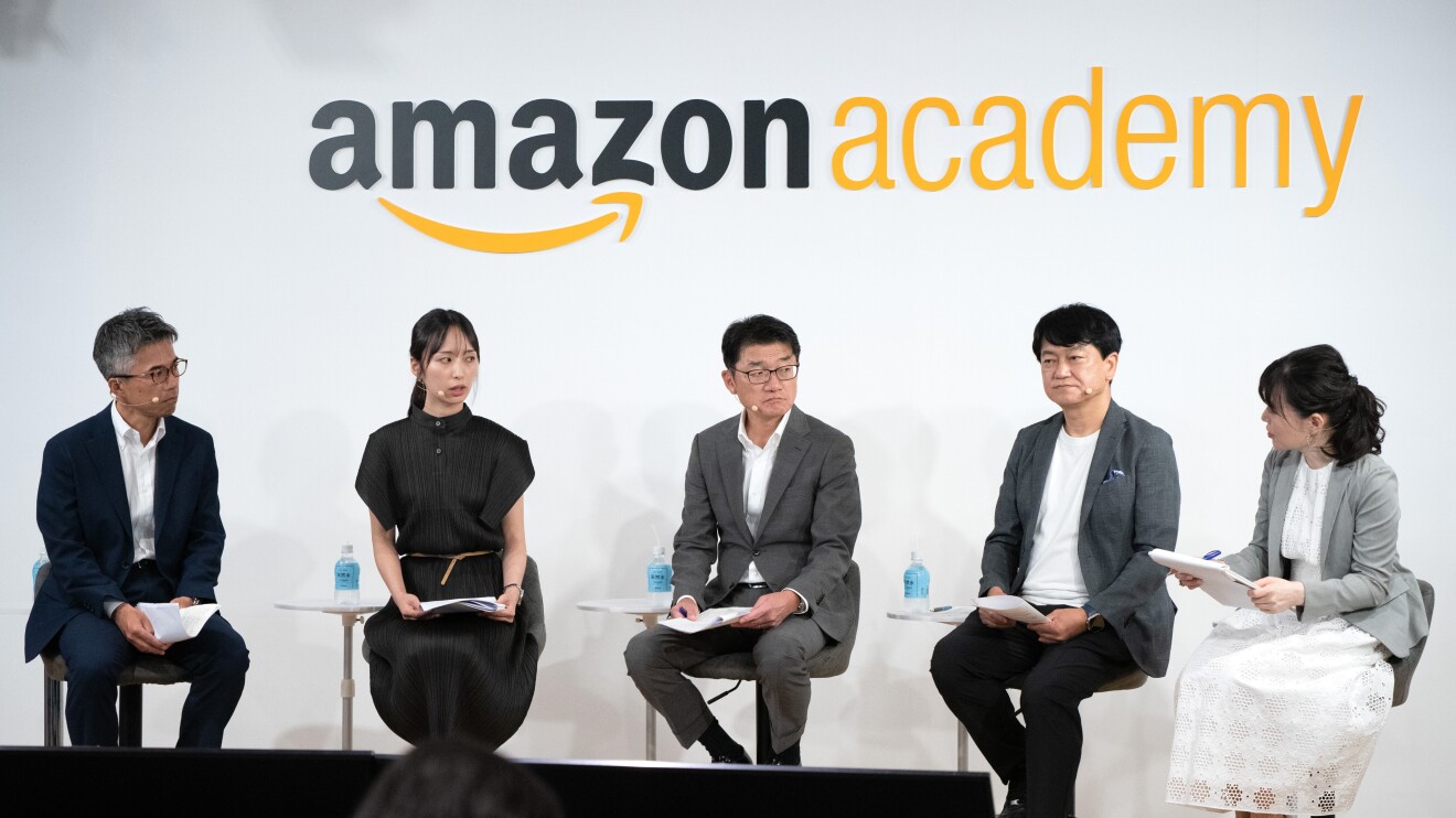 モノクローム服を着ている男性3人と女性2人が話し合いながら座って並んでいる。上には「Amazon Academy」のロゴが壁に貼られている。