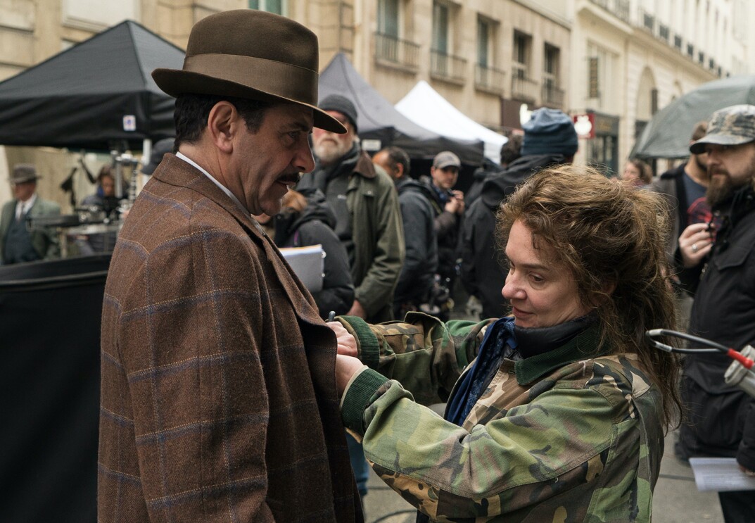 An image of a woman fixing a part of an actor's costume. They are standing in the middle of a filming set in a city street.