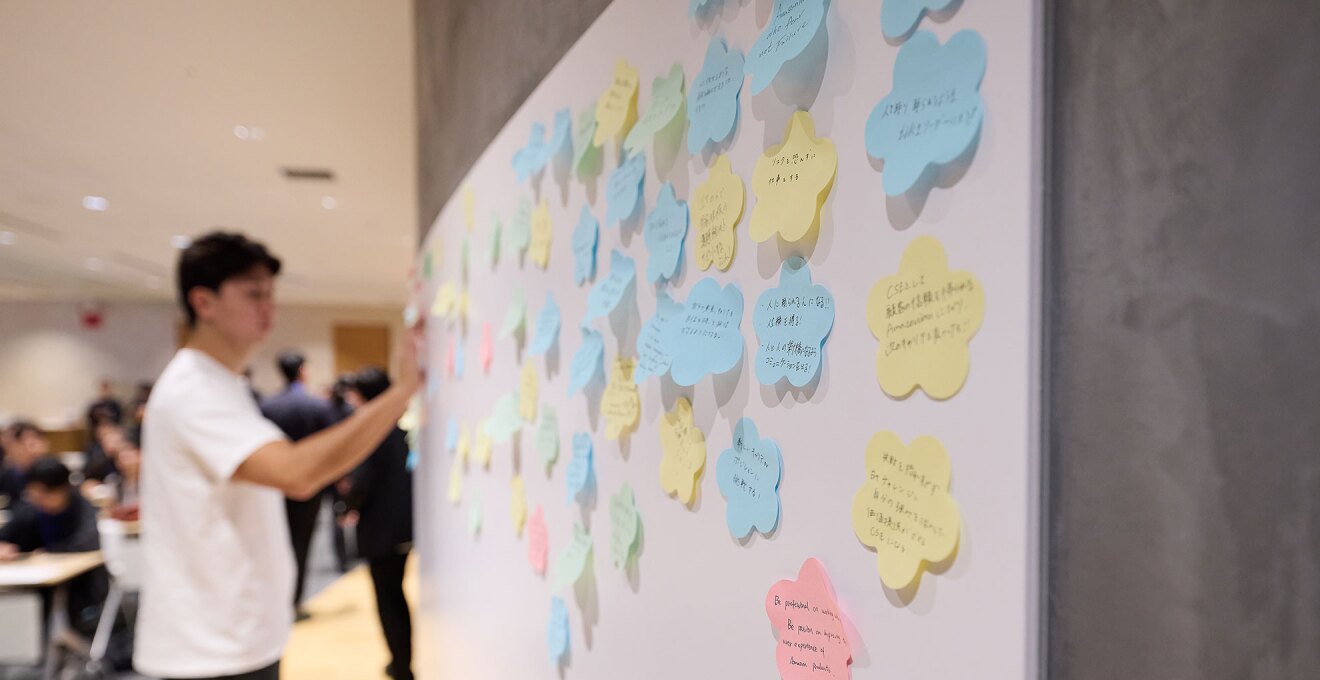 壁に貼られたホワイトボードに付箋を貼る若い男性。ボードには4色の花形の付箋が多数貼られている。