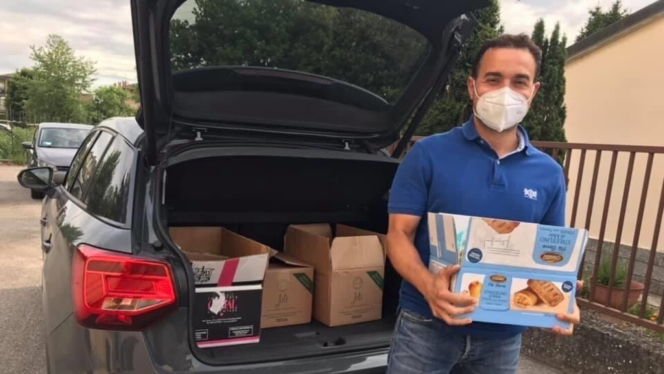 Un uomo davanti al baule di un'auto pieno di pacchi e viveri. Indossa una mascherina sul volto e regge con le mani un pacco di viveri. 