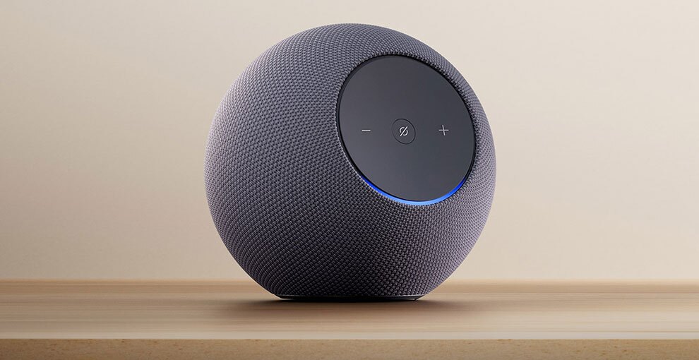ベージュの壁を背景に、木目調の台の上に置かれた球形の黒いデバイス。前面の丸いパネルに「-」「+」ボタンと、中央に斜線入りの丸アイコン。パネルの下側に青い光。パネル以外の本体表面はメッシュ状。