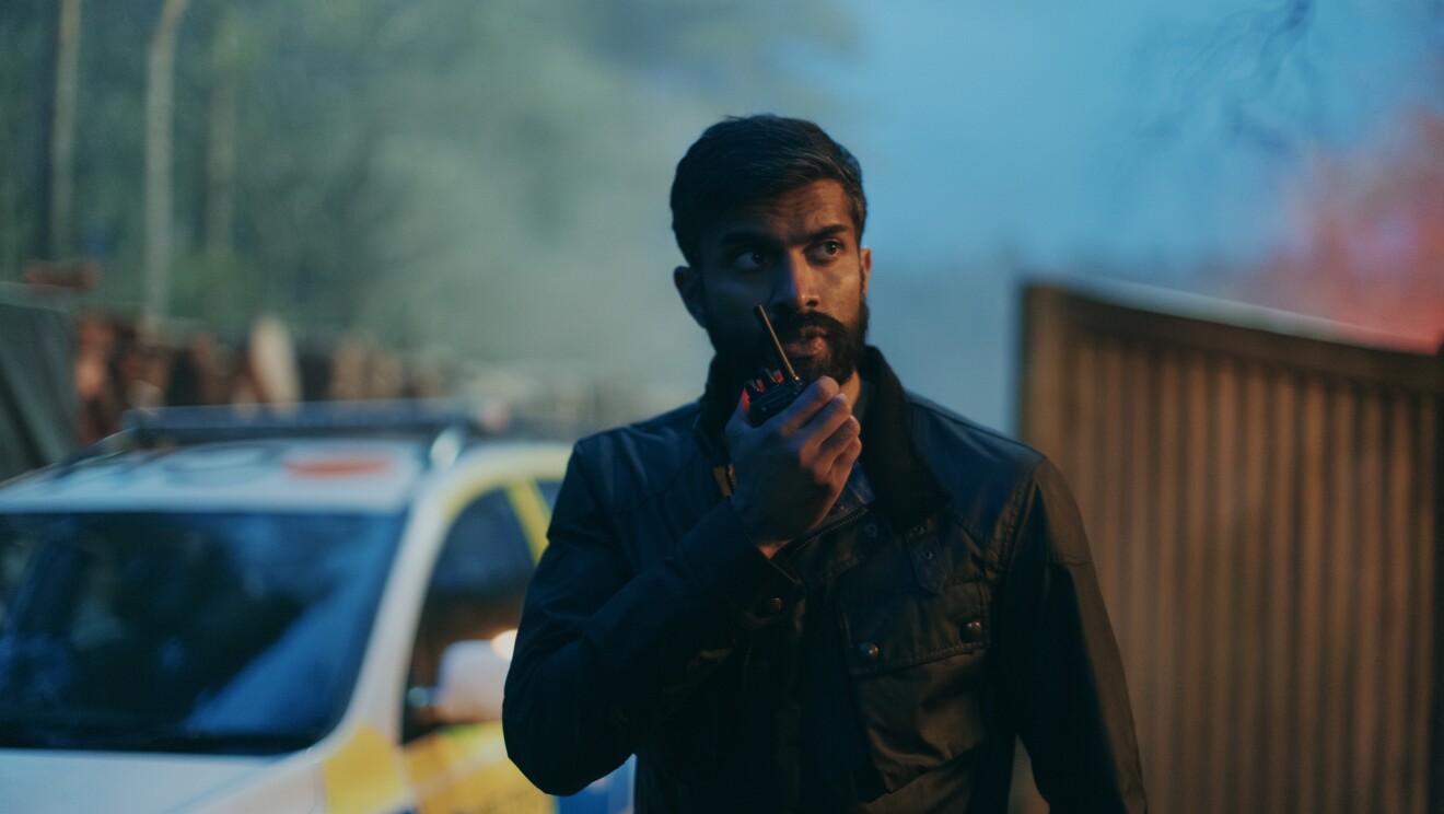 A bearded police officer holding a walkie talkie and  standing next to a police car