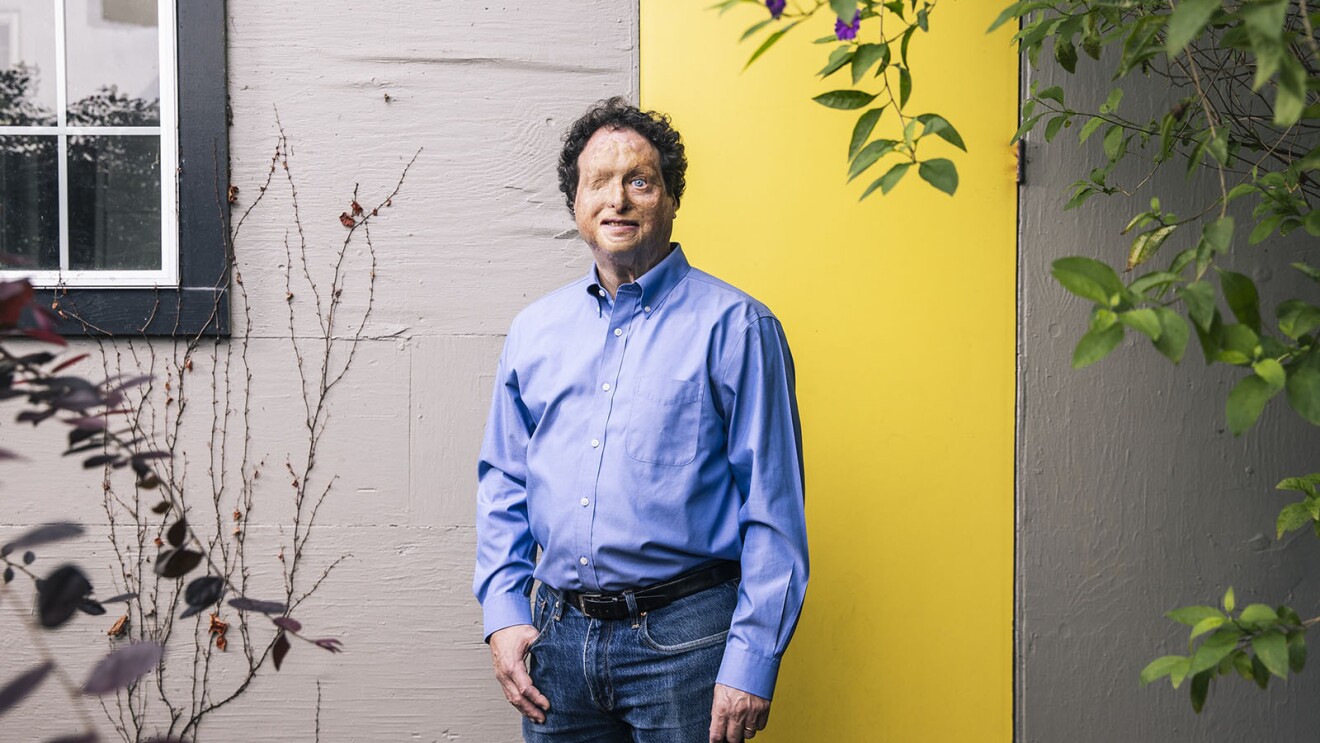 亚马逊126实验室的首席无障碍研究员Josh Miele在他位于加州伯克利的家中站着拍摄肖像。他站在家外面,在一堵浅灰色的墙前,有一扇小窗户和一扇亮黄色的门。在他的两侧有一棵小树和一株小植物。