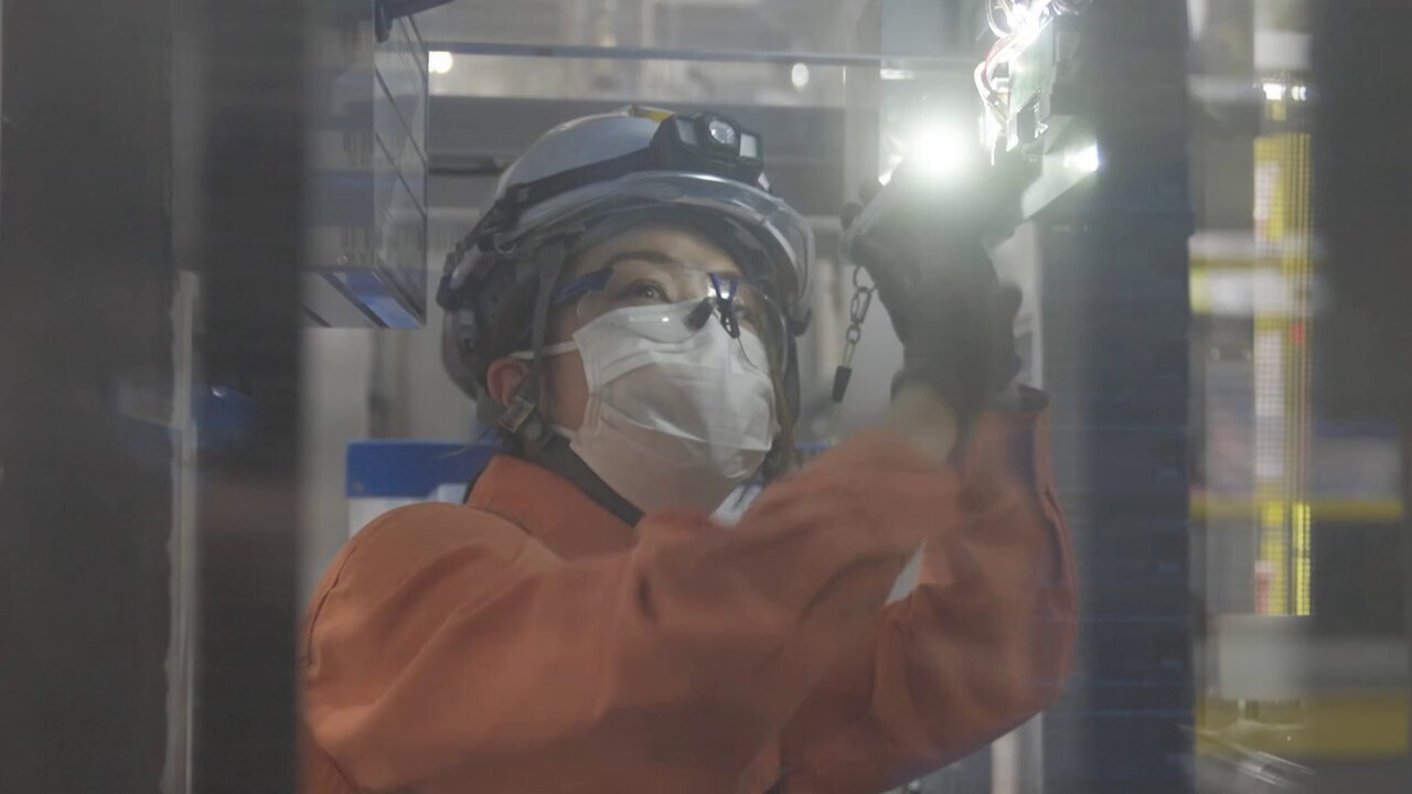 オレンジ色の作業着と白いヘルメット姿の女性。薄暗い中、懐中電灯で機械を照らしている