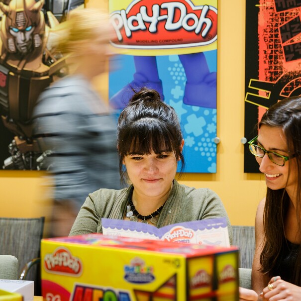 两个女人在前台坐在一张桌子周围包装。别人在后台经过。墙上有海报的小马驹,橡皮泥和其他孩之宝品牌。