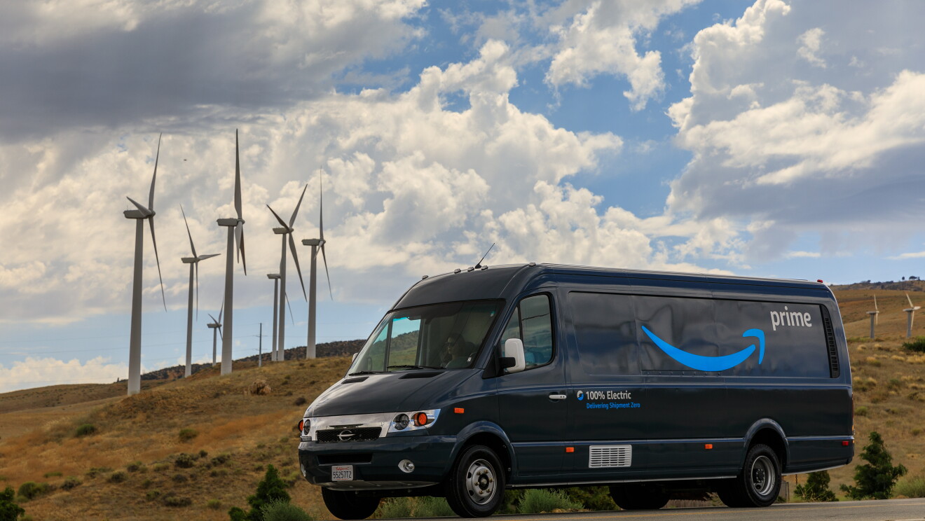  An Amazon van with "Prime," the Amazon Smile, and "100% electric" displayed on the side. The van is parked in front of several wind turbines. 