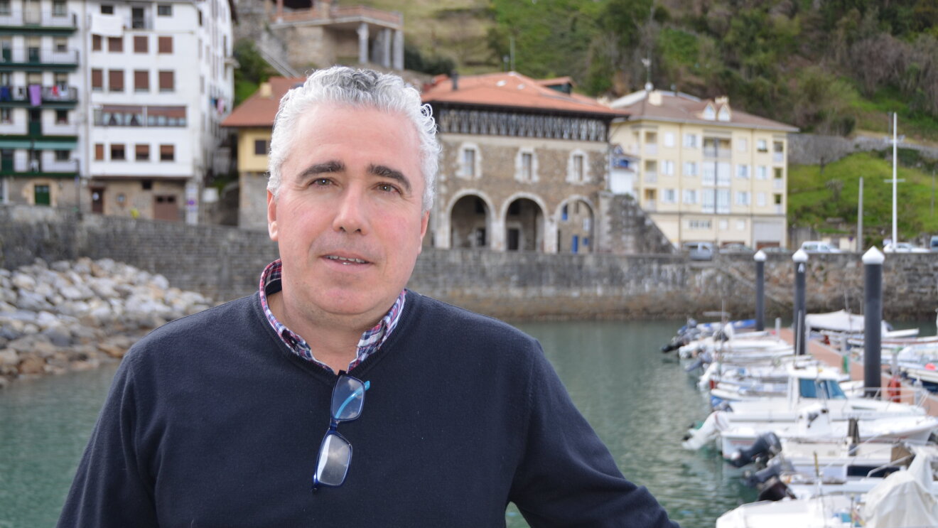 Juan Yurrita en el puerto de Mutriku. Luce un jersey azul con una camisa de cuadros y de fondo tiene las barcas del puerto y parte de las casas del pueblo.
