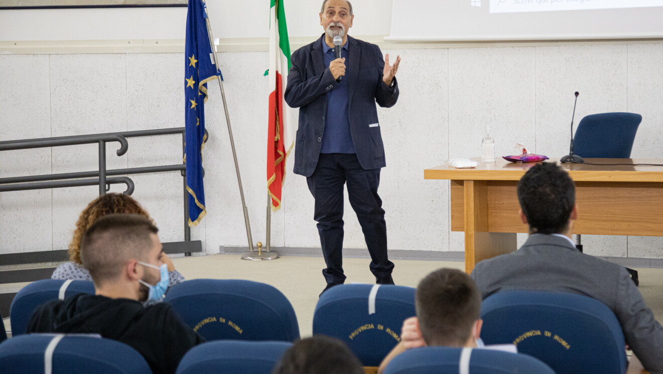 Foto di Umberto Guidoni mentre parla con gli studenti. Guidoni tiene un microfono nella mano destra, ha la barba bianca. Alle sue spalle vediamo una bandiera dell'Europa e una dell'Italia. 