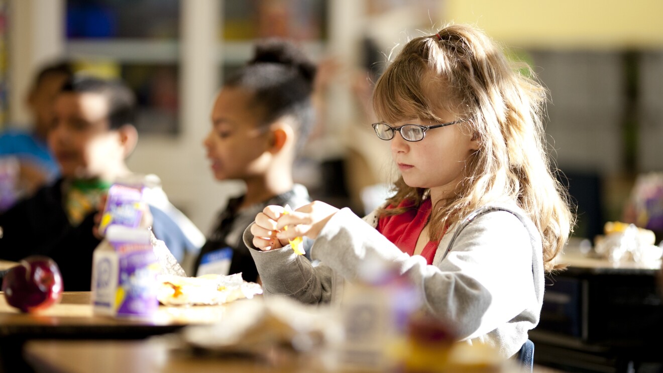 A young girl is opening preparing to eat. Behind her, other students are also eating. Apples and single-serve milk containers are on the desks in front of the students.