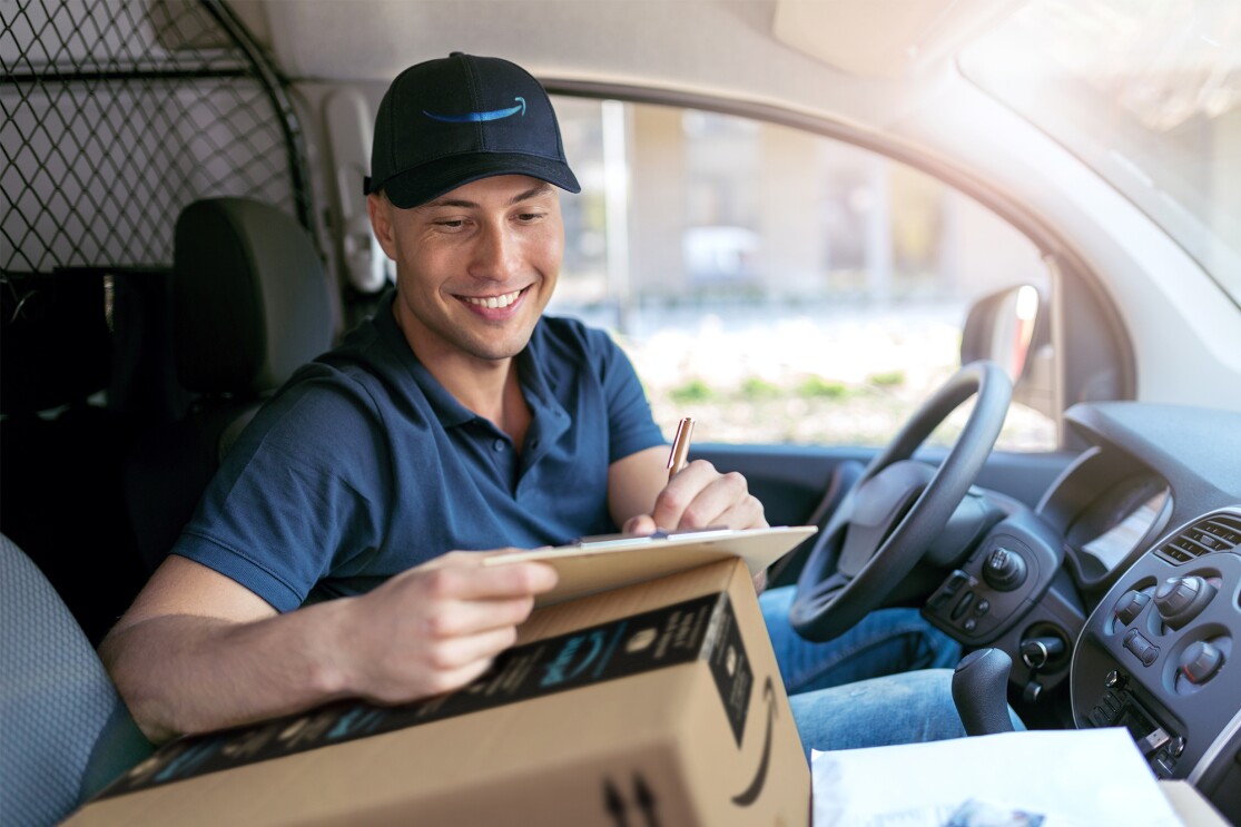 Un repartido de Amazon en una furgoneta. Está sentado en el asiento del conductor. Lleva un polo azul y una gorra de Amazon con la sonrisa en azul. Se está apoyando sobre una caja para escribir en una hora.