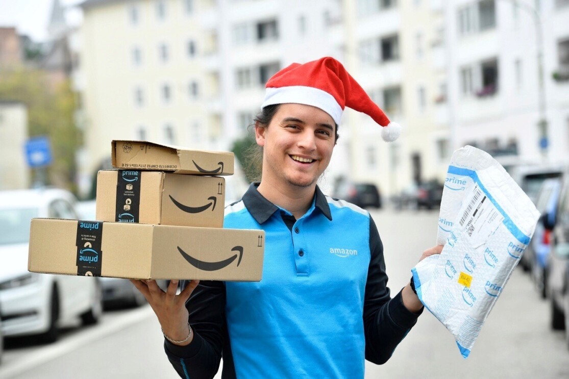 Ein junger Mann in Amazon Zustellkleidung und roter Zipfelmütze hält eine Reihe von Paketen in der Hand