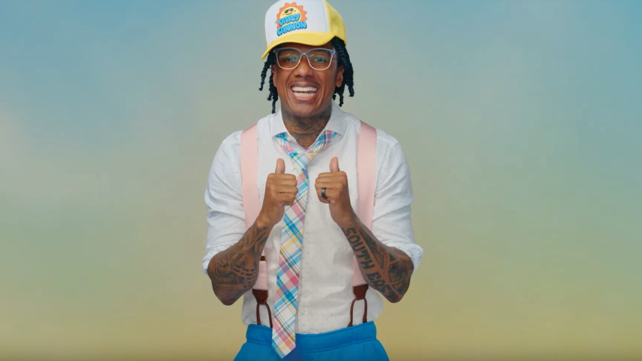Nick Cannon smiling to the camera with his two thumbs up and a blue and yellow ombre background. 