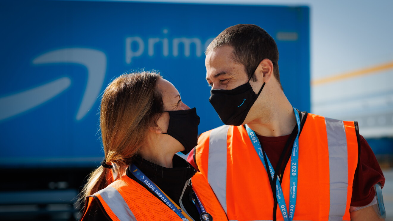 Sira Valeriano y Cristian Gamella. Foto de medio plano mirándose con mascarilla de color negro con la sonrisa azul de Amazon Es una foto exterior y de fondo se ve un trailer con la sonrisa de Amazon y la palabra Prime. Los dos protagonistas van con una camiseta negra, él de manga corta y ella una sudadera y él va con el pelo corto y ella largo recogido con una coleta.