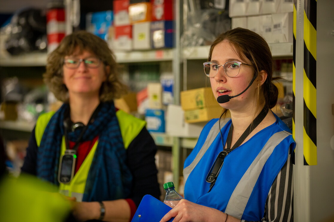 Im Bildvordergrund: Eine junge Frau mit Brille, Headset und blauer Weste. Links neben ihr (etwas unscharf) erkennt man eine Besucherin in gelber Sicherheitsweste.