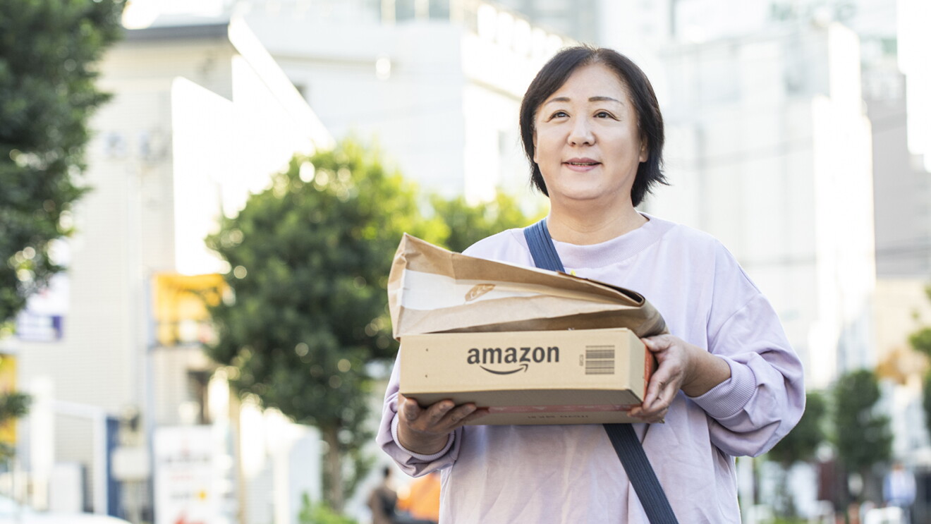 Amazonの段ボール箱と紙袋パッケージの2つを両手で体の前で持ち、街を歩く女性。背後に建物や街路樹が見える。