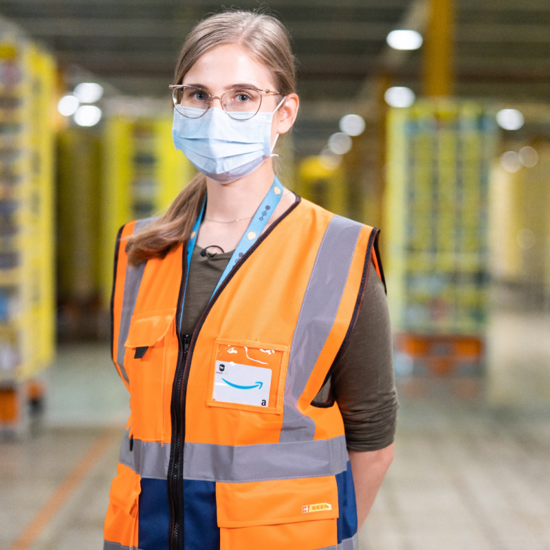 A woman wearing glasses, a face mask, and an orange safety vest stands in an Amazon fulfillment center. 