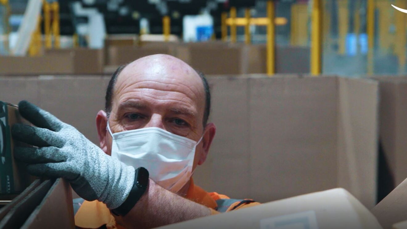 Primo piano di un dipendente Amazon mentre smista dei pacchi. Di fronte al suo mento la sua mano sinistra sta spostando un pacco su uno scaffale. Indossa sul volto una mascherina chirurgica. 