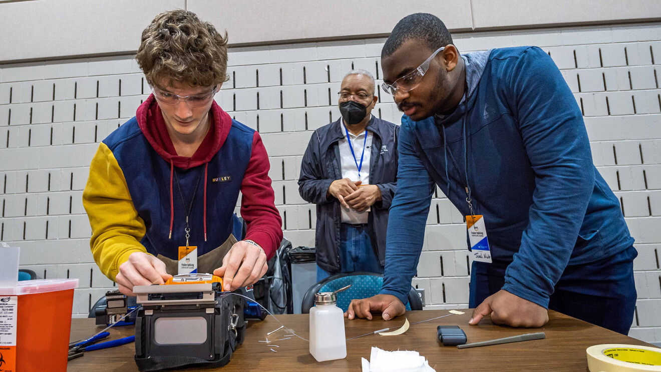 A photo of two students working on a fiber optic fusion splicer. An instructor is standing behind them.