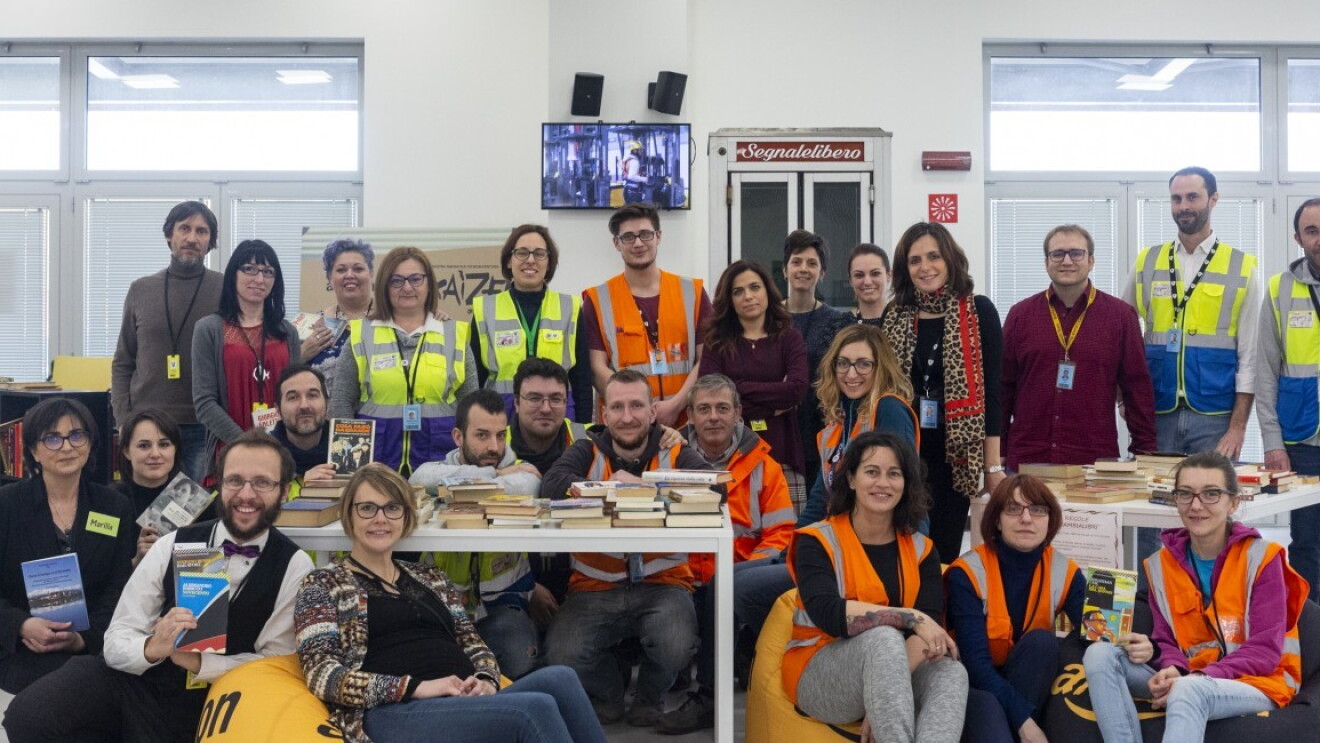 Un gruppo di donne e uomini riuniti in posa in un ufficio. Alcuni siedono su pouf morbidi, altri stanno in piedi. Alcune donne e uomini indossano dei giubbini gialli e arancioni di sicurezza. Tutti guardano verso la camera sorridendo. 