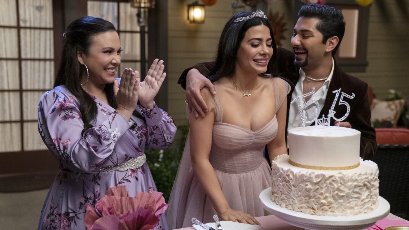  Gloria Calderón Kellett, Emeraude Toubia, and Mark Indelicato are celebrating the "double quincenera" of Emeraude's character, Lily, in their backyard. They are all cheering happily while looking down at the cake.