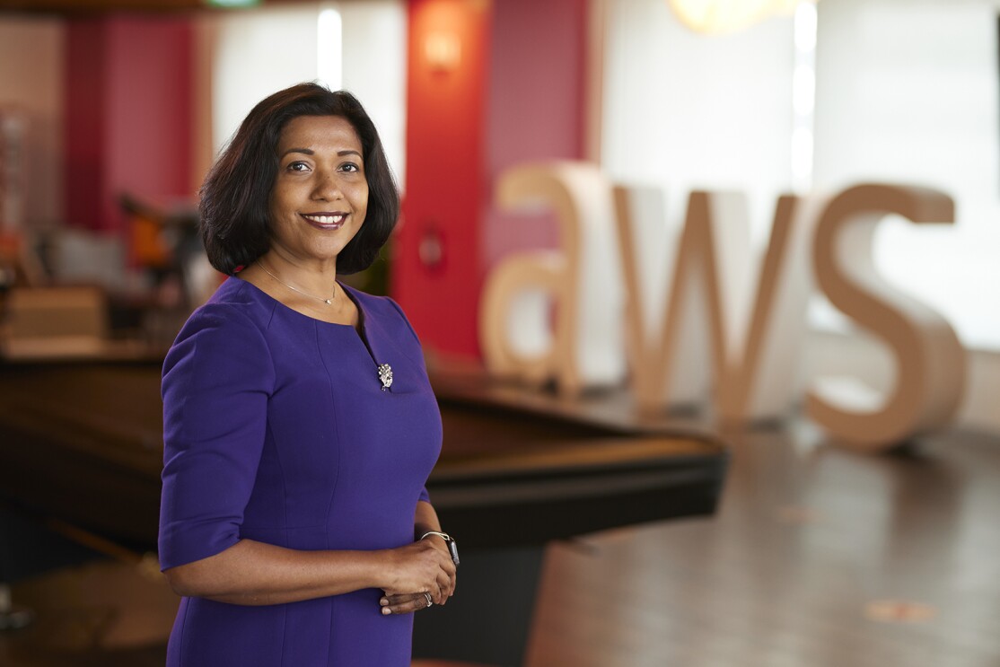 An image of a woman standing and smiling for a photo in a game room at an Amazon office. There is a large, letter sign in the background that says "AWS."