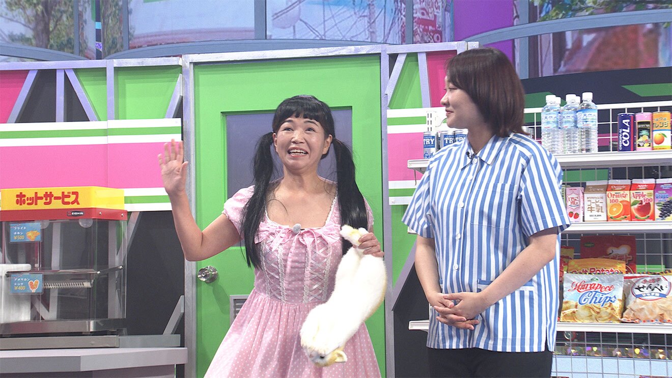 壁やドアが黄緑とピンクに塗られたコンビニを模したセットに、2人の女性が立つ。左側はピンクのワンピースに黒髪ツインテールでぬいぐるみを持ち、右側は白地に青ストライプのコンビニの制服。