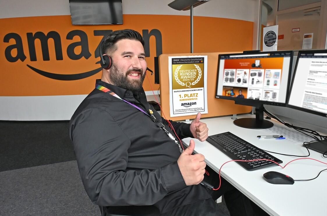 Deniz, Teamleiter beim Amazon Kundenservice an seinem Arbeitsplatz im Berliner Amazon Kundenservice Zentrum, mit Daumen-hoch-Geste: Er ist in schwarz gekleidet, trägt Kopfhörer und sitzt an einem weißen Schreibtisch mit schwarzer Tastatur und 2 Bildschirmen. An der Seitewand seinen Arbeitsplatzes hängt das Siegel des Deutschen Kundenservice Awars 2024/25.