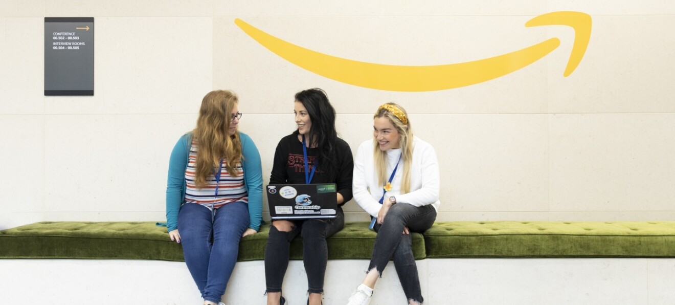 Drei junge Frauen sitzen auf einem Sofa. Sie haben einen Laptop in der Hand. Im Hintergrund sieht man das Amazon Logo.
