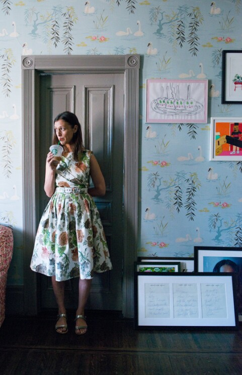 An image of Sally Singer standing in a room with blue wallpaper and interesting art on the walls. She is sipping a cold drink from a Starbucks cup and looking out the window.