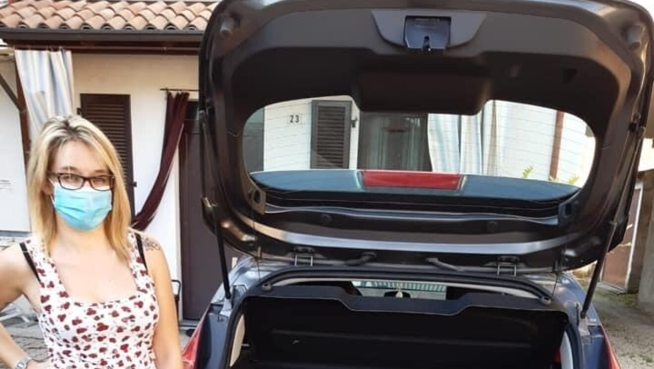 Una donna davanti al baule di un'auto pieno. Indossa una mascherina sul volto e guarda verso la camera. 