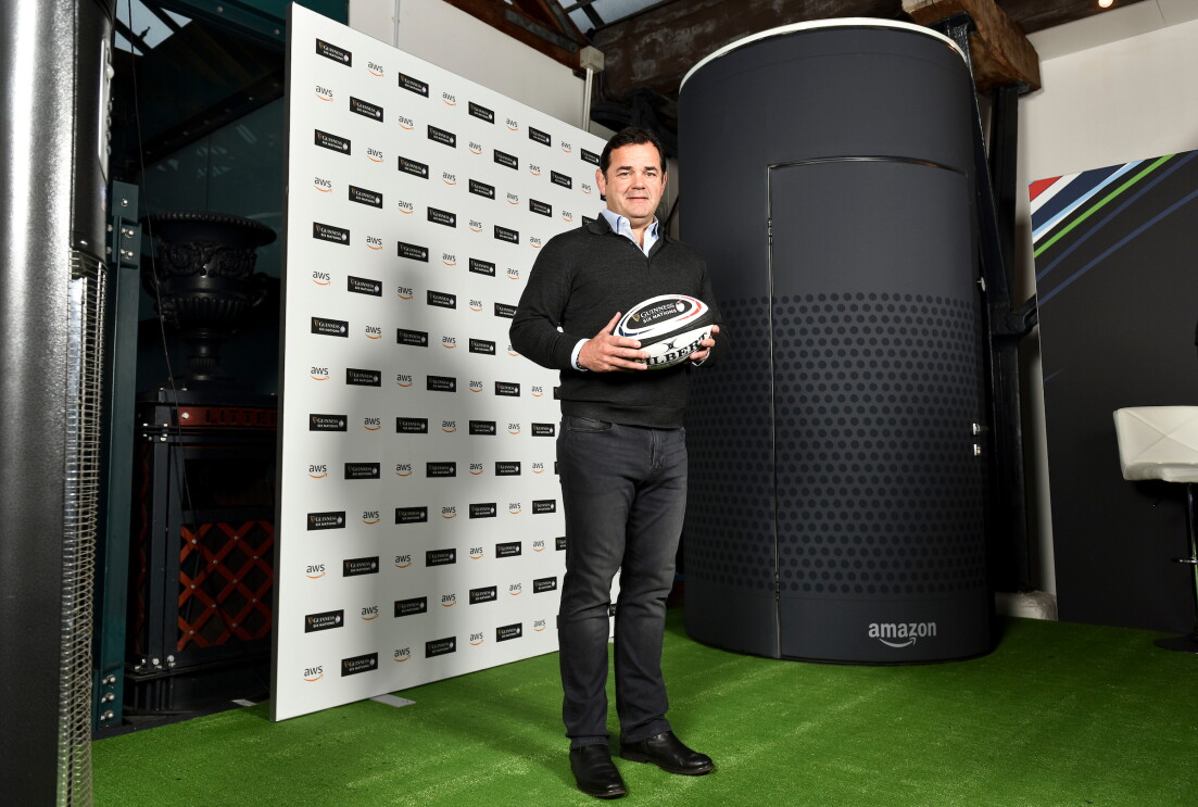 Will Carling standing holding a rugby ball at the Six Nations promoting AWS. He is standing in front of a large Amazon Alexa Echo model.