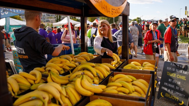 社区香蕉站的前景这张照片所示,与与会者成堆的香蕉,免费参加由两个“bananistas。”In the background, event attendees are seen walking and standing.