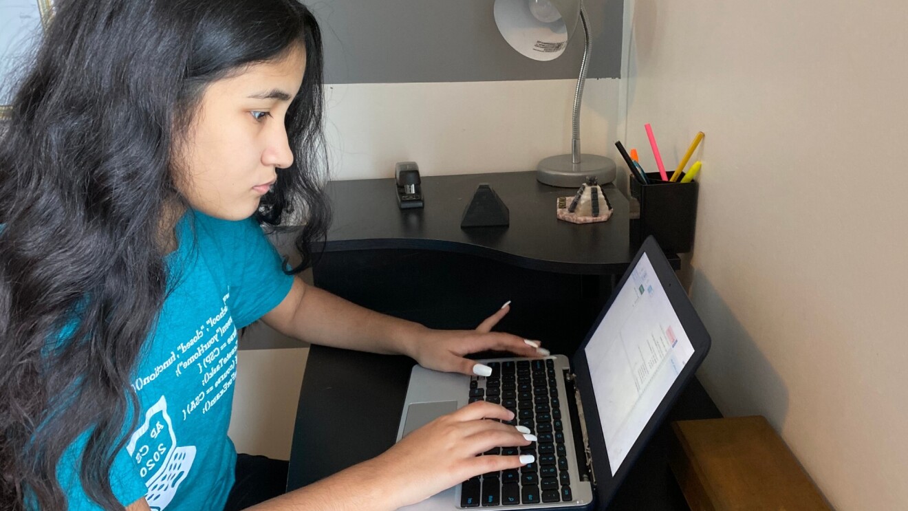 A young woman types on a laptop at a desk in her home. She wears a blue t-shirt from a coding program.